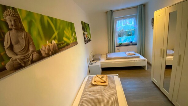Una habitación pequeña y limpia con una cama, un banco de meditación, una estatua y una ventana con vistas.