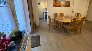 Un comedor con una mesa redonda y seis sillas, una ventana con cortinas de encaje y una cocina al fondo.