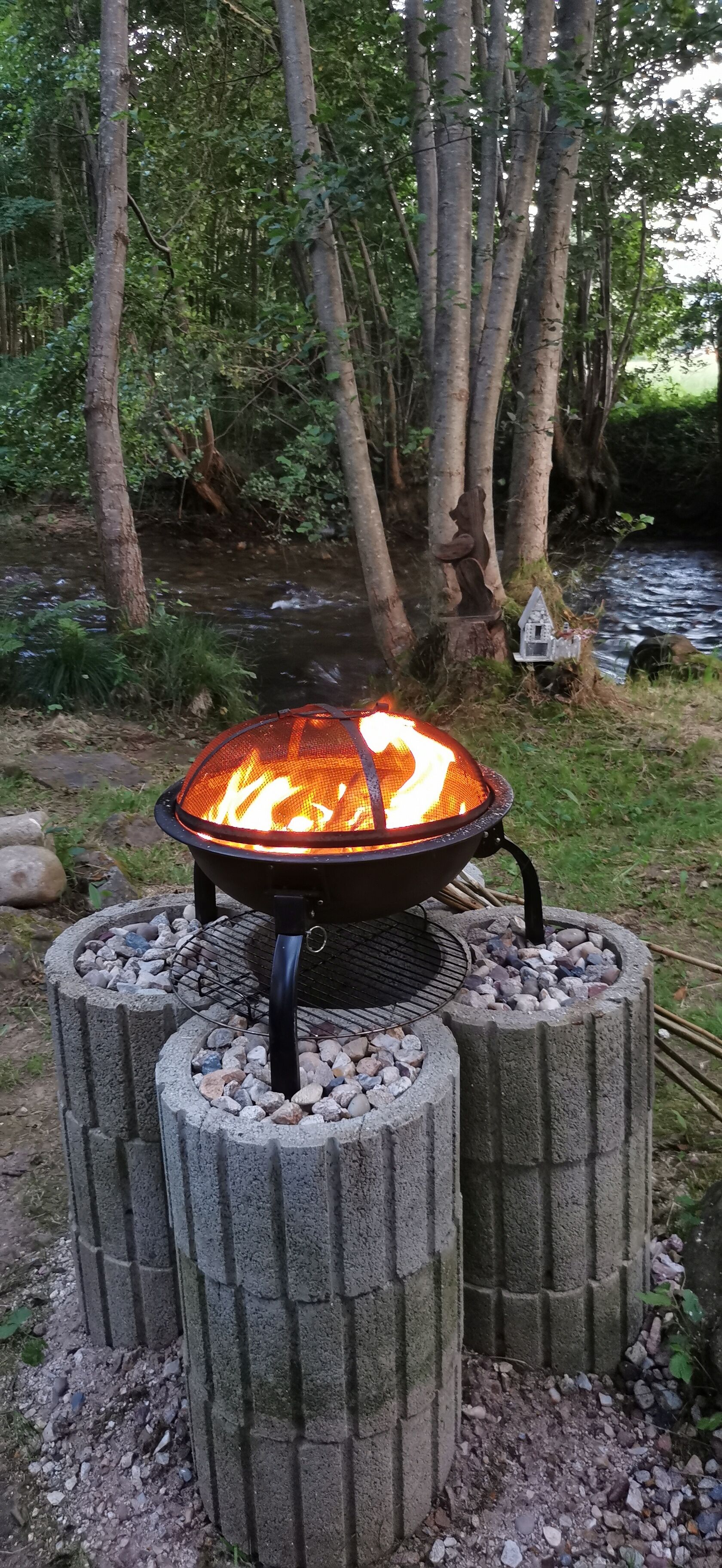 A portable fire pit with a visible flame, surrounded by stone supports, set in a natural outdoor environment with trees and a stream in the background.