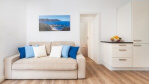 Un dormitorio con un sofá beige, cojines azules y blancos, suelo de madera, armario blanco y una encimera de cocina con fruta.