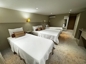 Une chambre d’hôtel avec trois lits simples, une petite table de chevet, une lampe et un appareil de climatisation fixé au mur.