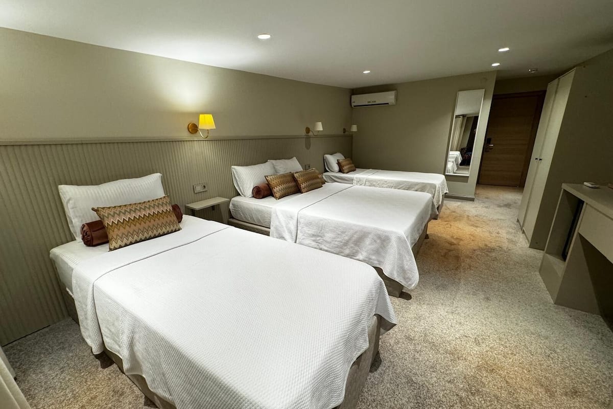 A hotel room with three single beds, a small bedside table, a lamp, and an air conditioning unit on the wall.