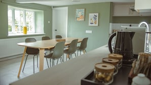 A dining area with a wooden table and chairs, a window with a view, and a kitchen area with a kettle and jars. - Kestrel Cottage in Castle Ashby (Northampton)