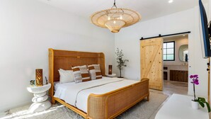 A spacious bedroom with a large bed, a wooden headboard, a rattan-framed bed, a white bedside table, a wall-mounted TV, and a sliding door leading to a bathroom. - Casa De Tierra - Featured in Architectural Digest (Palm Springs)
