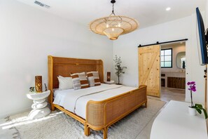 Un dormitorio amplio con una cama grande, cabecera de madera, estructura de ratán, mesita de noche blanca, televisor montado en la pared y una puerta corredera que da a un baño.