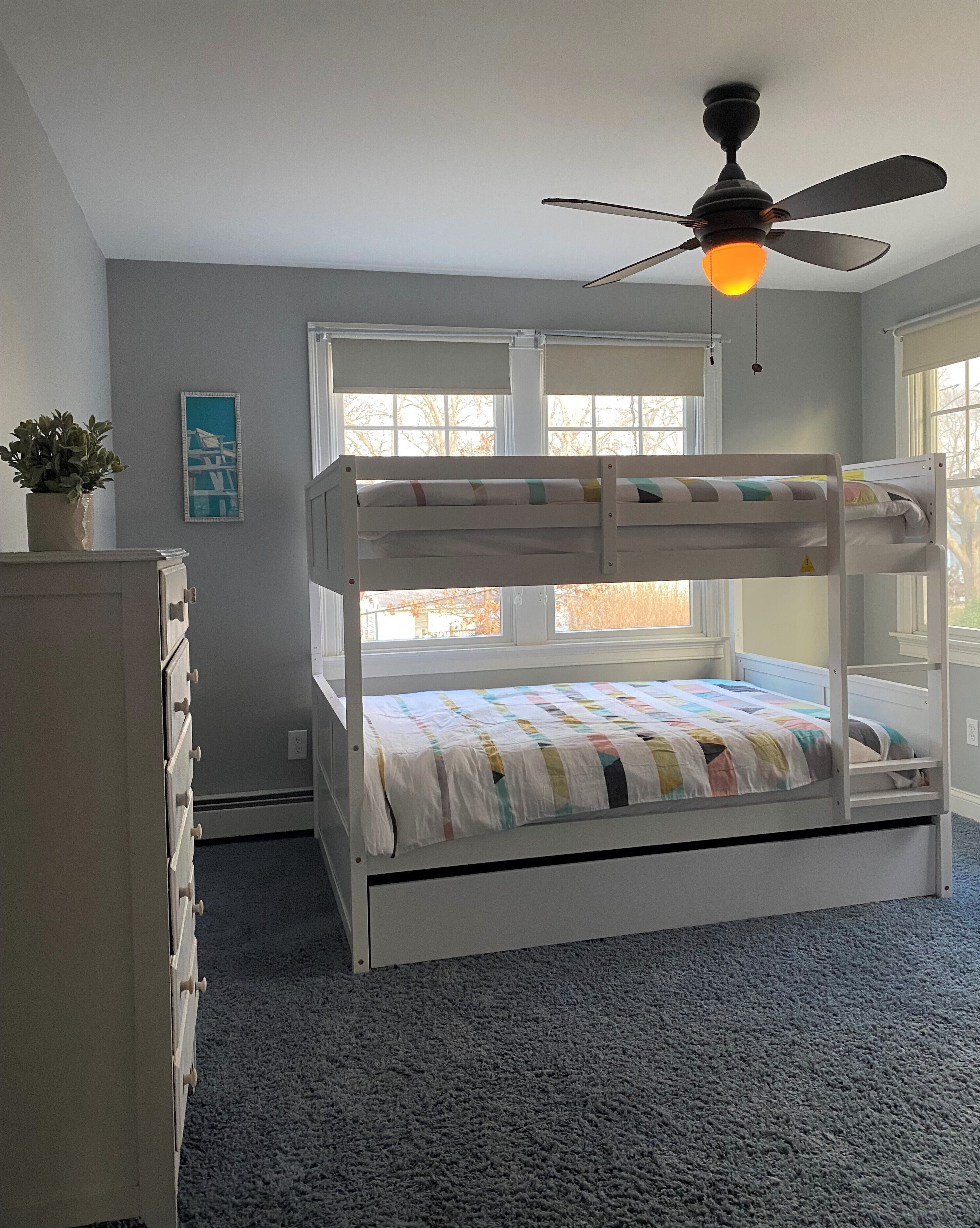 Una cama litera con ventilador de techo, cómoda, planta y ventana con vista a árboles.