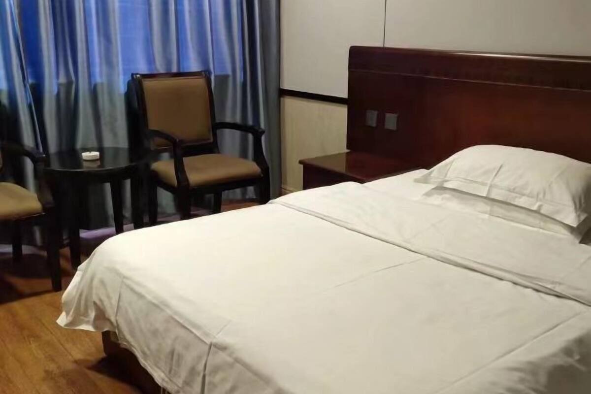 A hotel room with a large bed, a wooden headboard, a small table with chairs, and a view of the outside through curtains.