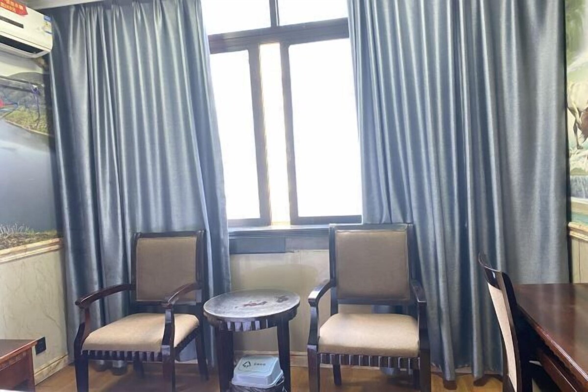Una habitación con dos sillas de madera y una pequeña mesa redonda, una ventana con cortinas, y un aire acondicionado instalado en la pared.