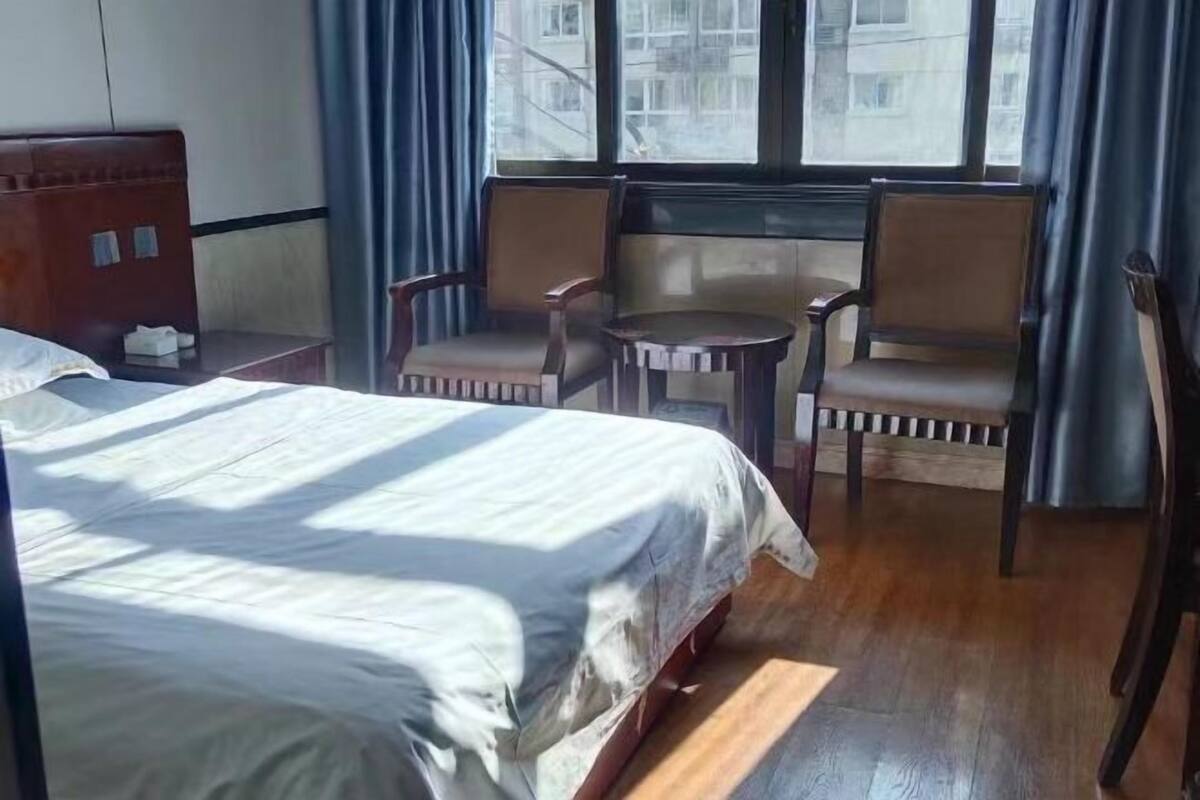 A hotel room with a large bed, two armchairs, a small table, and a view of buildings through a window.