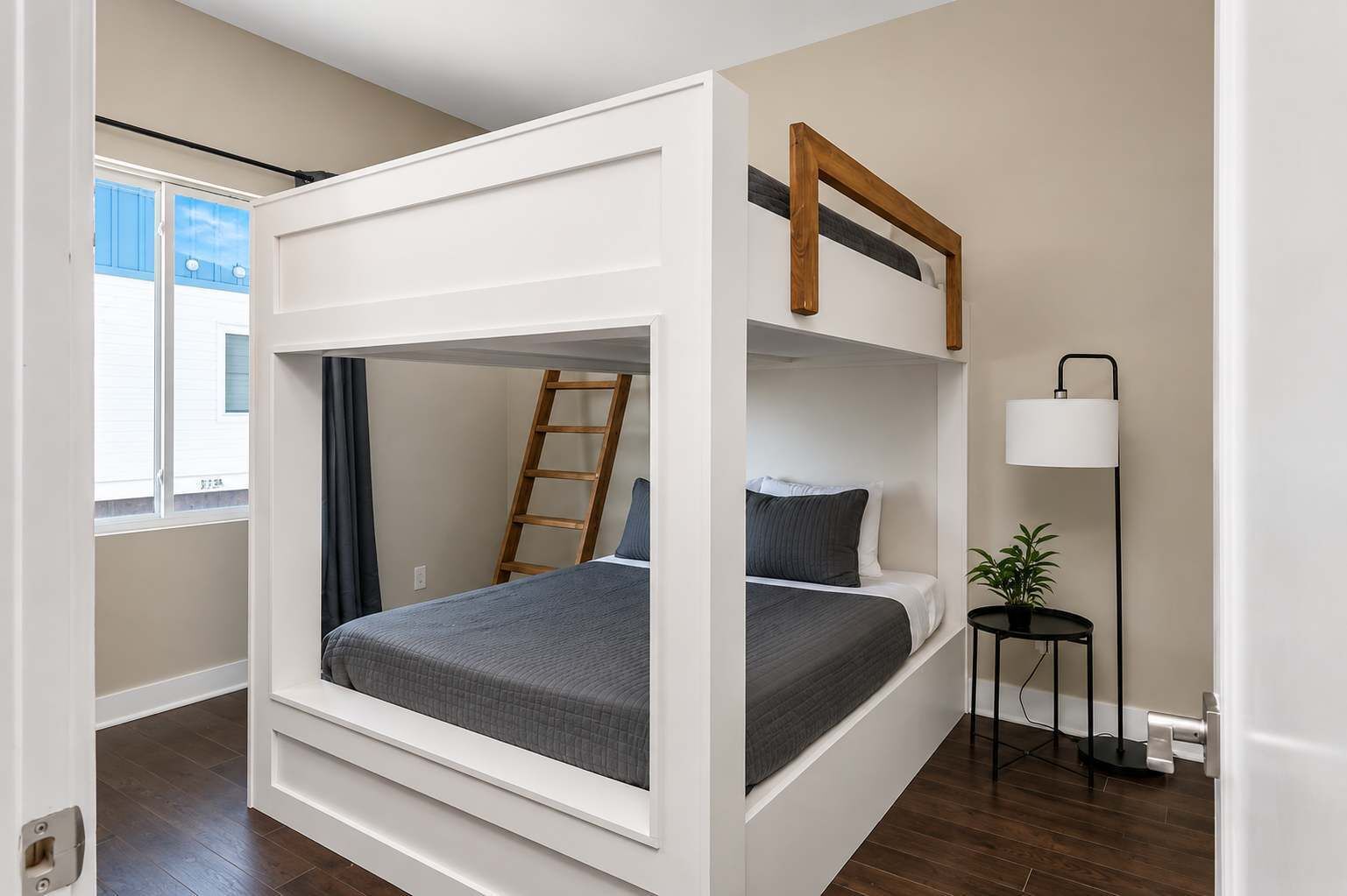 A bunk bed room with a ladder, a bedside table, a lamp, and a window with curtains.