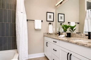 Ein modernes Badezimmer mit einer Badewanne, einer Duschvorhang, einem Waschtisch mit Waschbecken und einem Spiegel. Auf dem Waschtisch liegen Handtücher und ein Blumentopf.
