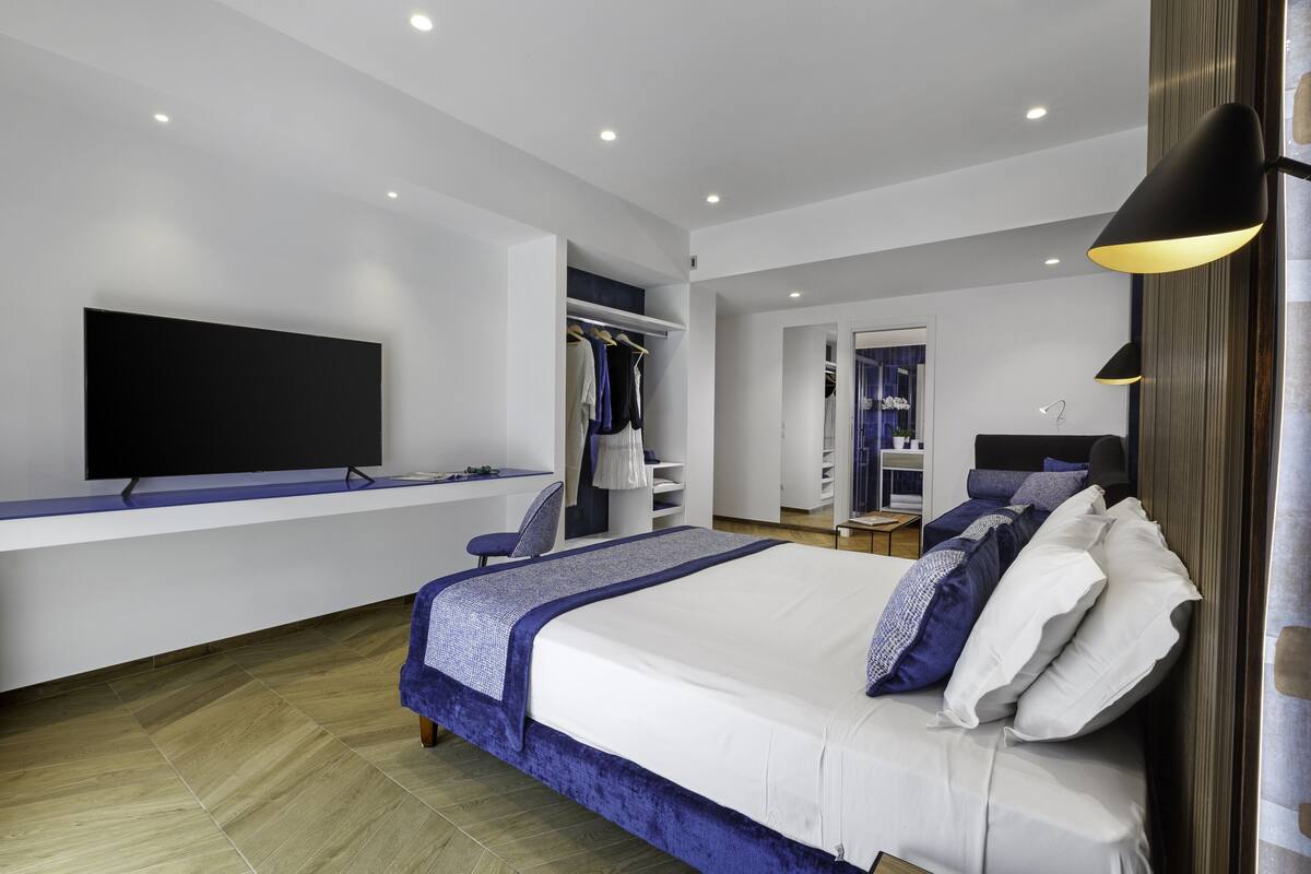 A modern bedroom with a large bed, a flat-screen TV, a desk, and a walk-in closet.