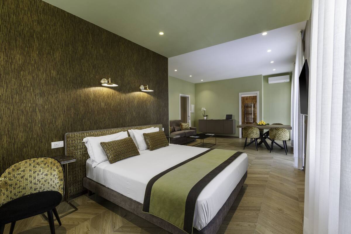 A hotel room with a large bed, a sitting area with a sofa and armchairs, a dining table with chairs, and a kitchen area in the background.
