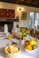 Un desayuno con una mesa lista para dos personas, con fruteros, un jarrón de flores y un cesto de pan.