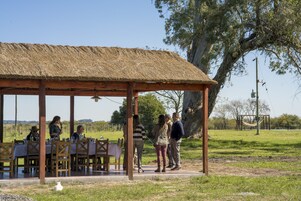 Un área de comedor al aire libre de estilo rústico con mesas y sillas de madera, una estructura de techo de paja y un campo de césped al fondo.