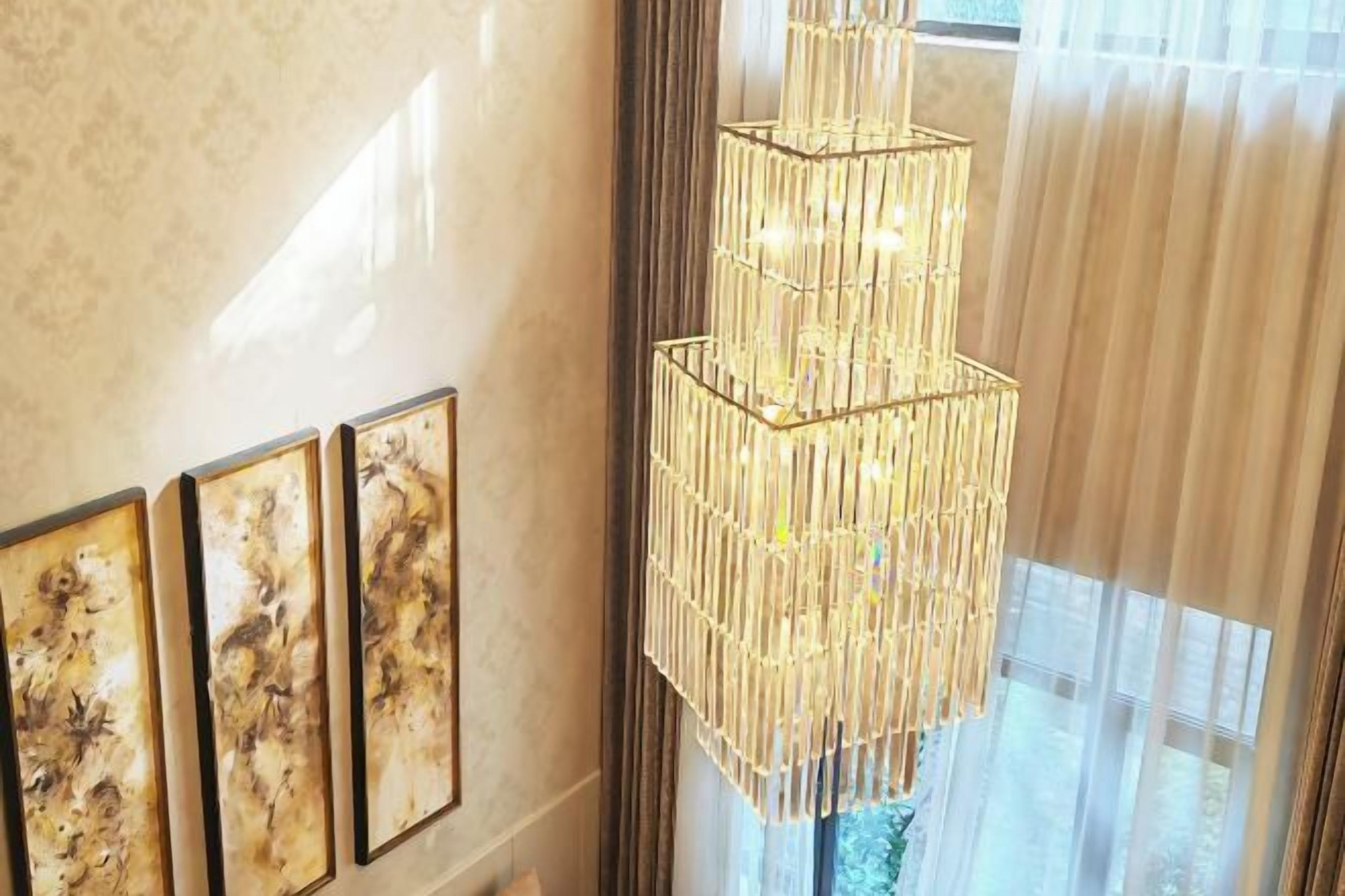 Un lustre au design géométrique est suspendu dans une pièce dotée d’une grande fenêtre et de trois œuvres d’art encadrées accrochées au mur.