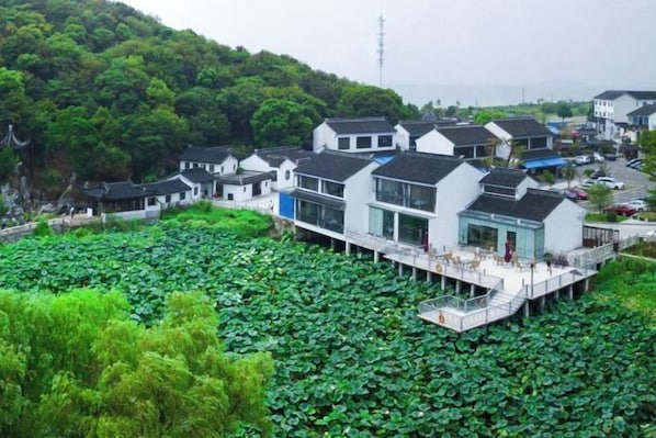 A traditional-style building with a modern extension, situated on stilts over a body of water with lush greenery around. - Juanshui Hostel (Suzhou)
