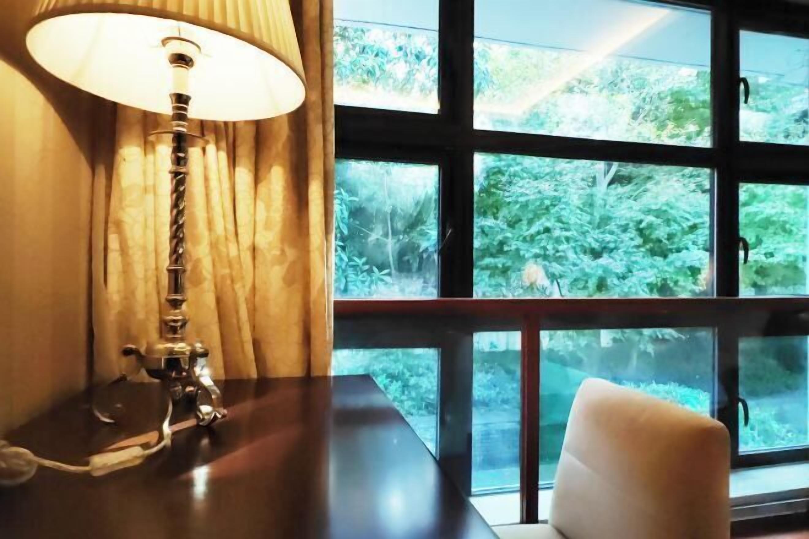 A room with a table, a lamp, and a window with a view of greenery.