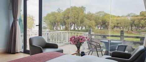 A hotel room with a large bed, two armchairs, a small table, and a balcony with a view of trees and a park.