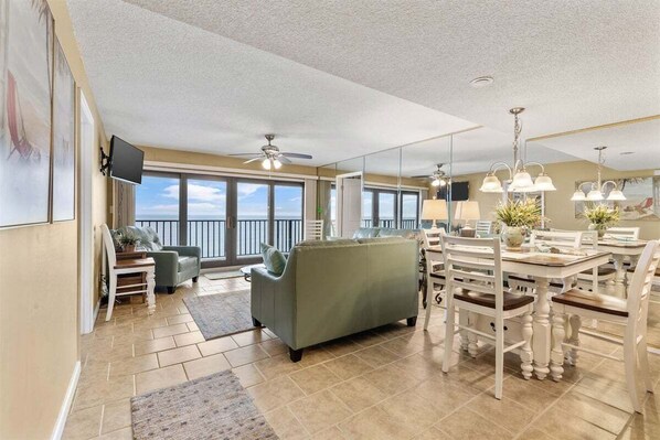 A spacious living area with a large window, a green sofa, a dining table with chairs, and a ceiling fan.