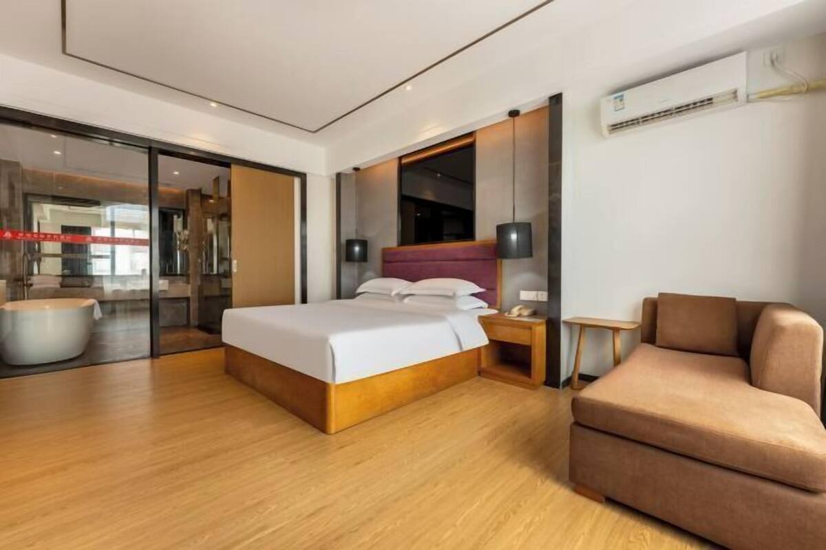 Une chambre d’hôtel moderne avec un plancher en bois, un lit avec du linge de maison blanc, un canapé, une table de chevet et un appareil de climatisation.
