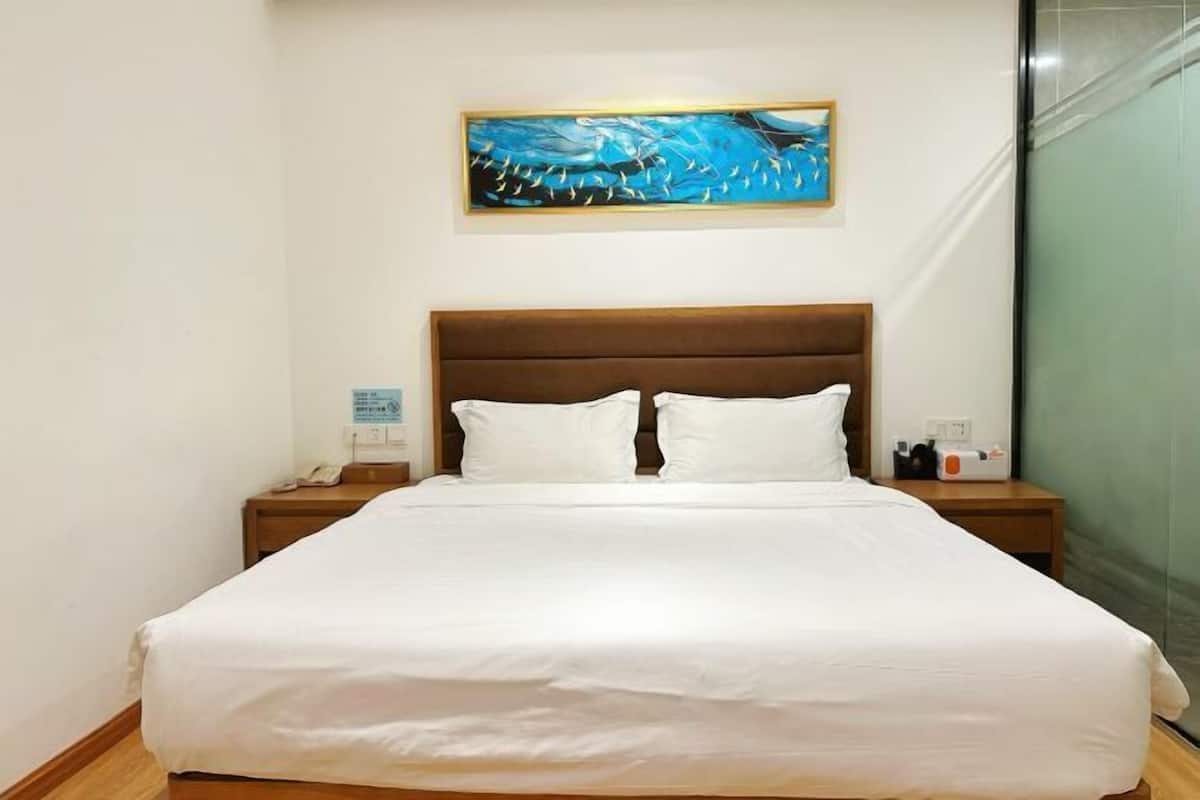 Une chambre d’hôtel avec un grand lit, une tête de lit en bois, du linge de lit blanc, un tableau au-dessus du lit, une table de chevet avec un téléphone et une lampe, et une porte de douche en verre.