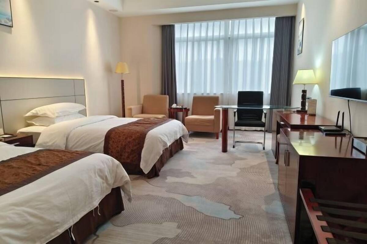 Habitación de hotel con dos camas, un escritorio, una silla, un televisor y un ventanal grande con cortinas.