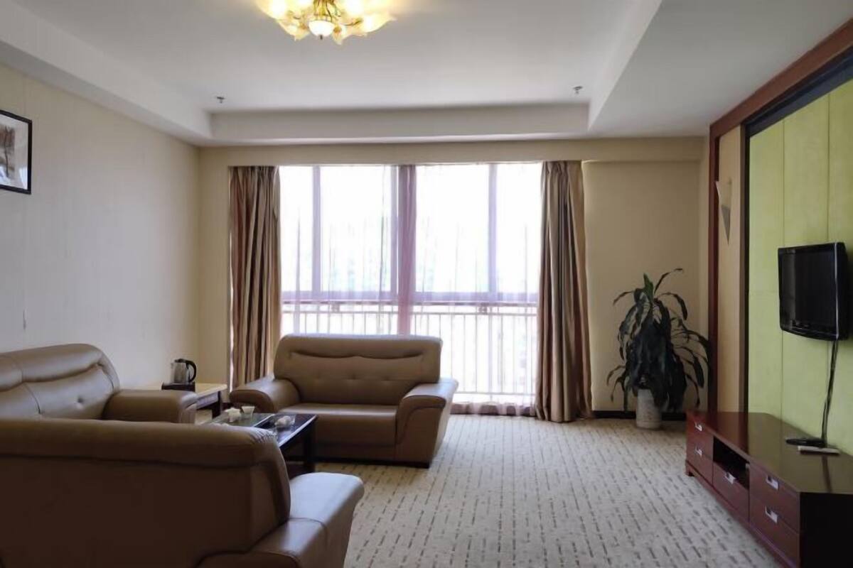 Una habitación de hotel con sofá, televisor, un ventanal con cortinas y una planta en maceta.