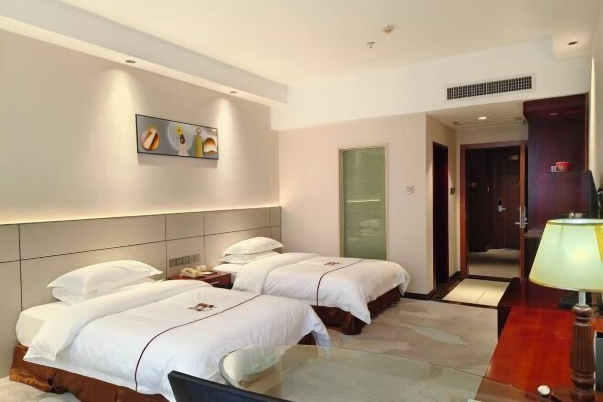 Habitación de hotel con dos camas, una mesita de noche, una lámpara, un cuadro en la pared y una puerta que da a otra habitación.