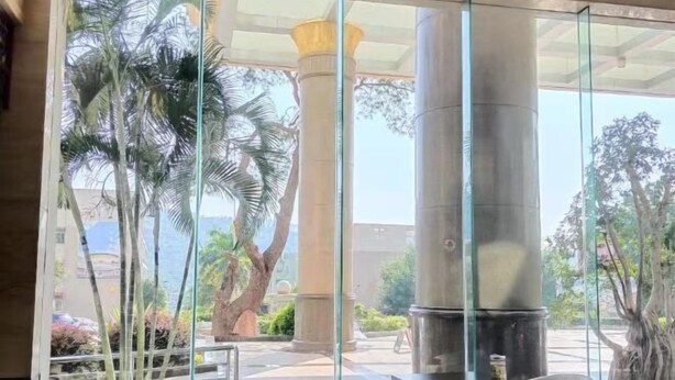 A restaurant with glass walls, outdoor seating, and a view of palm trees and a large column. - Hua'an Hotel (Hua'an)