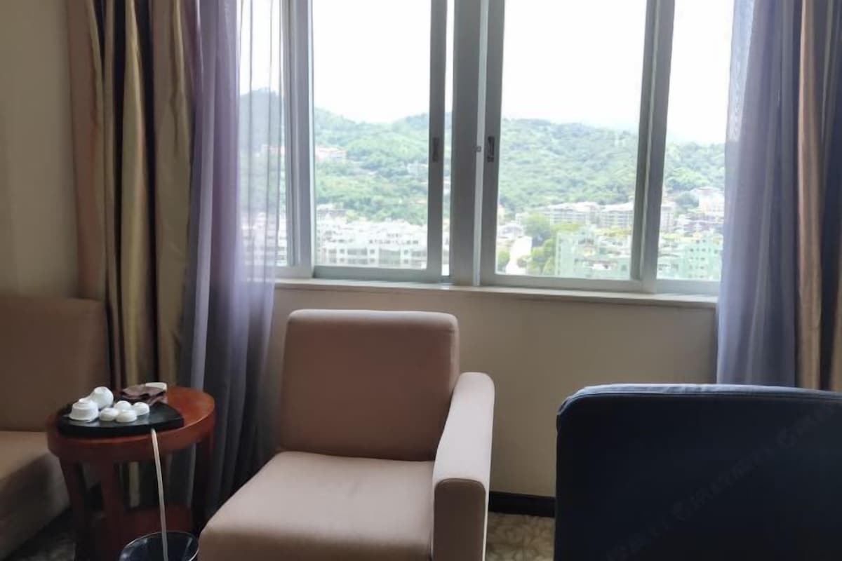Een hotelkamer met een beige fauteuil, een klein tafeltje met theepotten en uitzicht op een stadsgezicht door grote ramen.