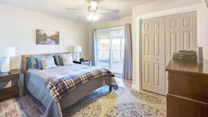 A bedroom with a bed, nightstands, a ceiling fan, a large window with curtains, and a door.