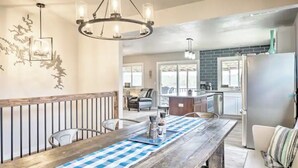 A dining area with a wooden table, chairs, and a blue and white checkered runner. There is a chandelier above the table and a kitchen in the background.