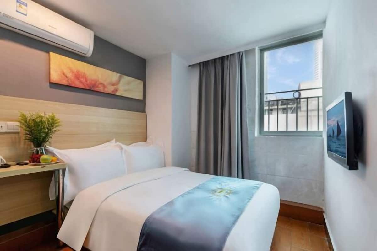 Ein Hotelzimmer mit einem Bett, einem Schreibtisch mit Pflanze, einer Klimaanlage, einem Fenster mit Vorhängen und einem an der Wand befestigten Fernseher.