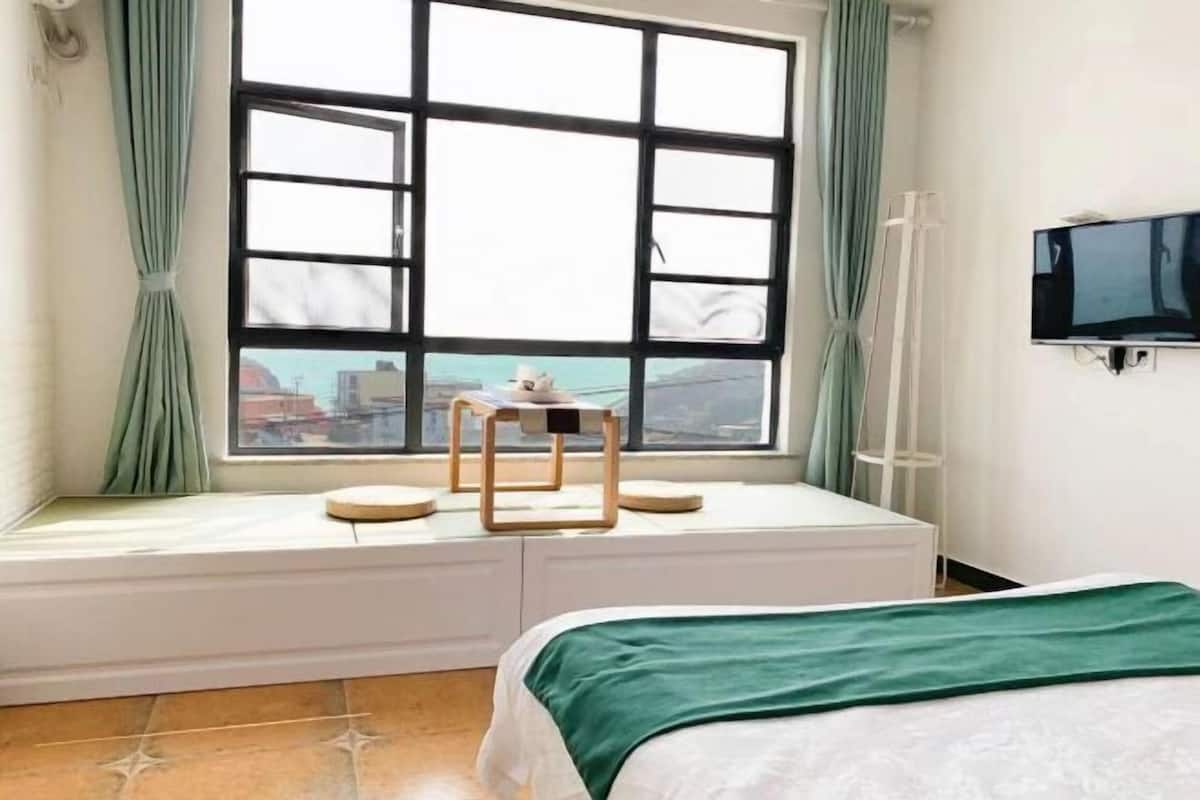 Un dormitorio con un ventanal grande, una cama, una mesita y un televisor instalado en la pared.