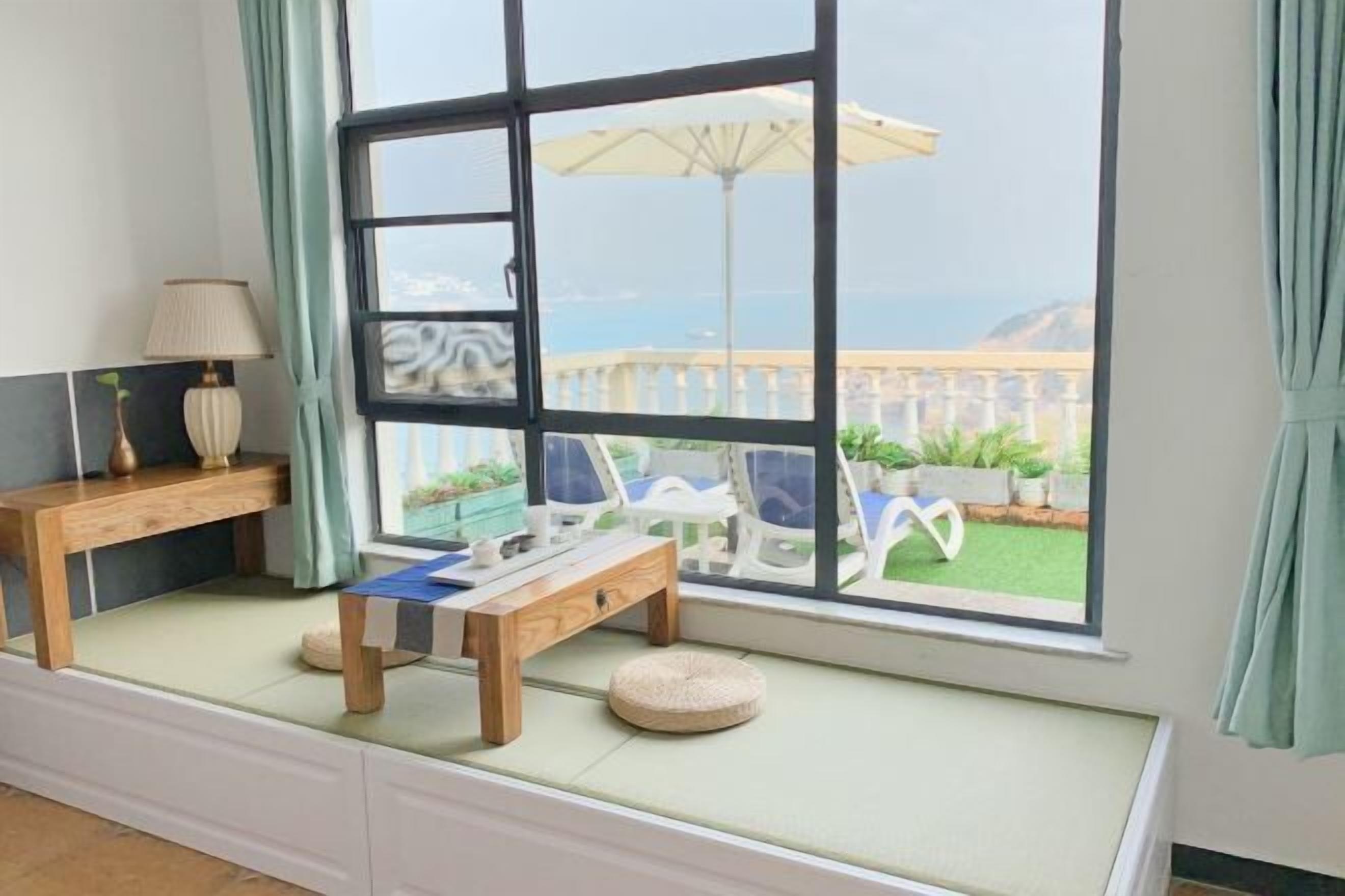 A room with a large window, a wooden bench, a small table, and a cushion.