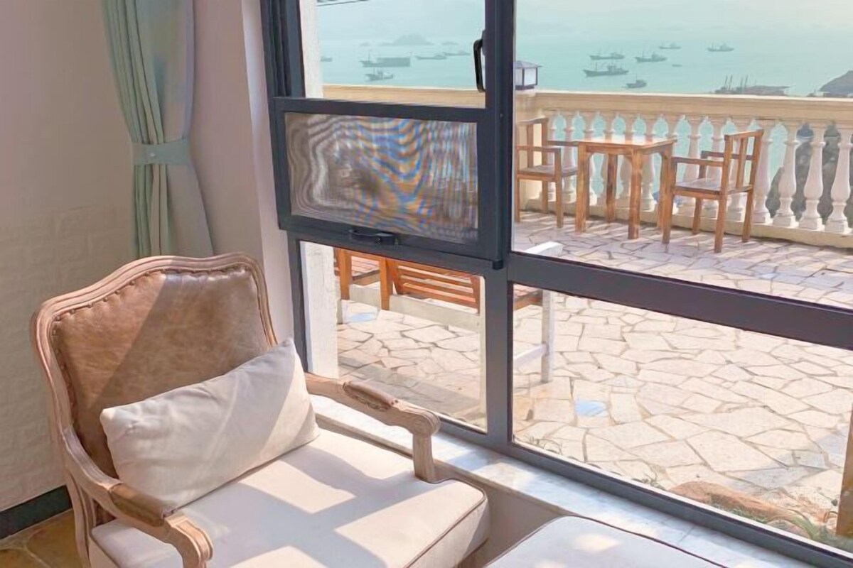 Une pièce dotée d’une grande fenêtre, d’un fauteuil confortable, d’une table en bois et d’un balcon avec une balustrade.