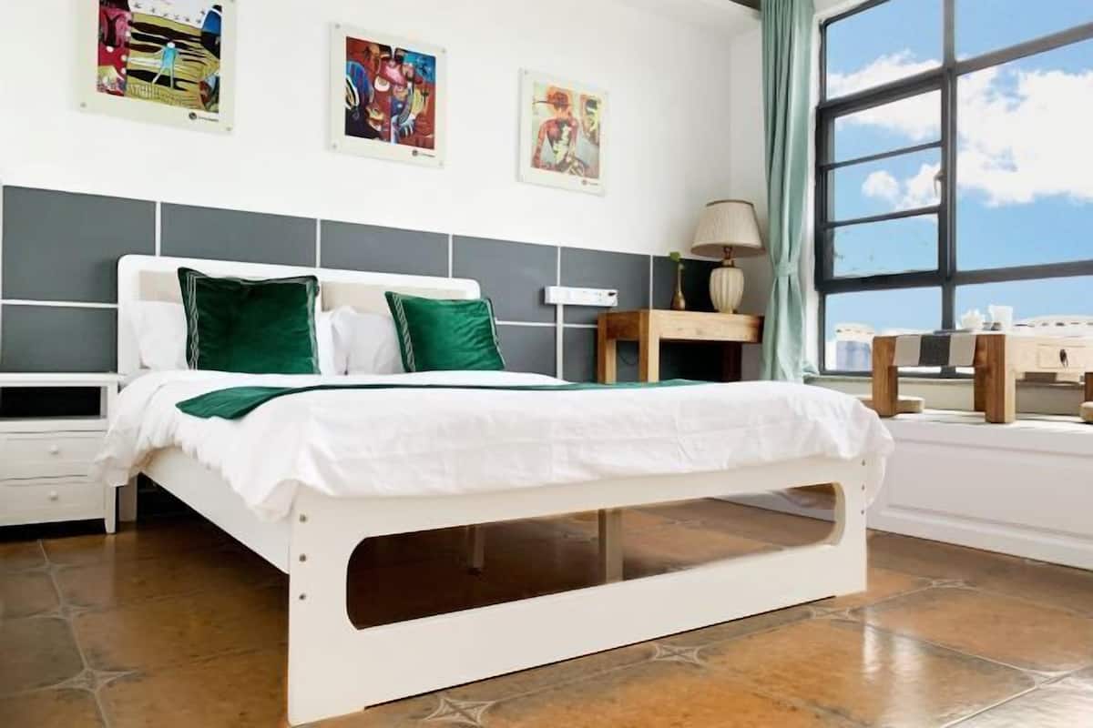 Una cama con estructura blanca y almohadas verdes, una mesita de noche, una lámpara y una ventana con vistas.