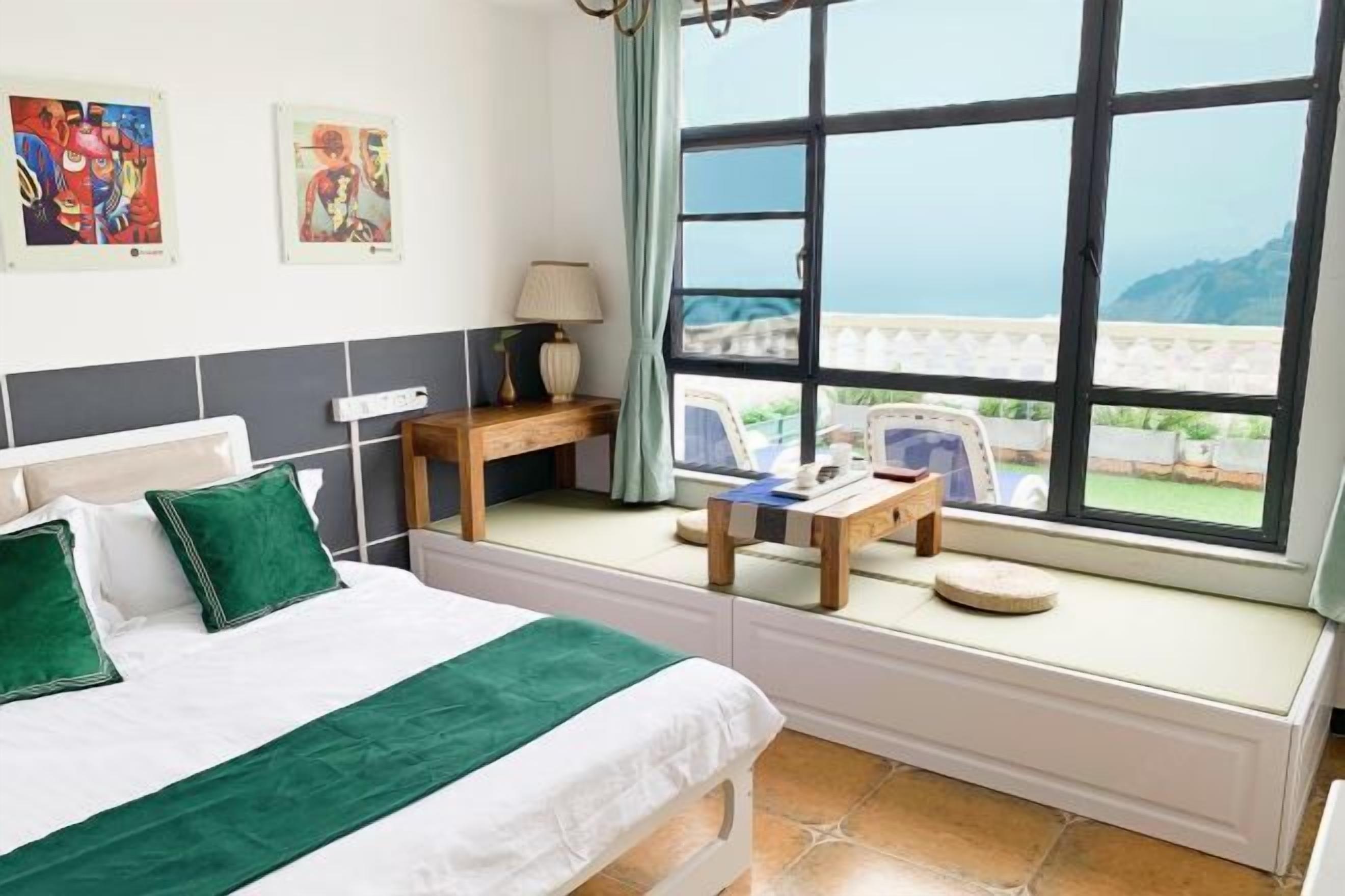 A hotel room with a bed, a small table, a chair, a window with a view of the sea, and artwork on the wall.