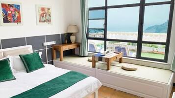 A hotel room with a bed, a small table, a chair, a window with a view of the sea, and artwork on the wall.