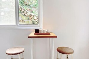Une pièce avec une fenêtre donnant sur un mur de pierre et sur la verdure, une petite table en bois sur laquelle se trouve un plateau avec des tasses et une théière, et deux tabourets dotés d’assises rembourrées.
