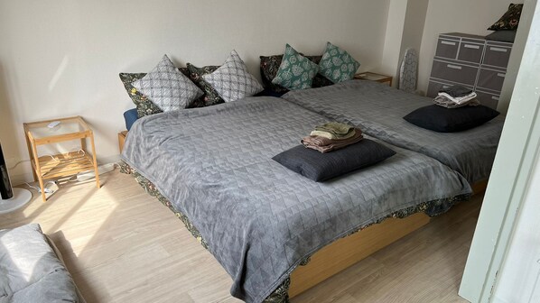 A neatly made bed with a grey quilt, multiple patterned pillows, and a small wooden side table with a power strip.