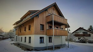 A two-story wooden house with a balcony, surrounded by snow-covered trees.