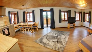 A spacious room with wooden floors, a dining area with chairs and a table, a television on a stand, and large windows with curtains.