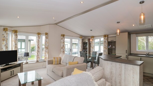 A modern living room with a TV, sofa, and glass coffee table. There is a kitchen area with a bar and dining space. - Gwydn Lodge (Lymington)