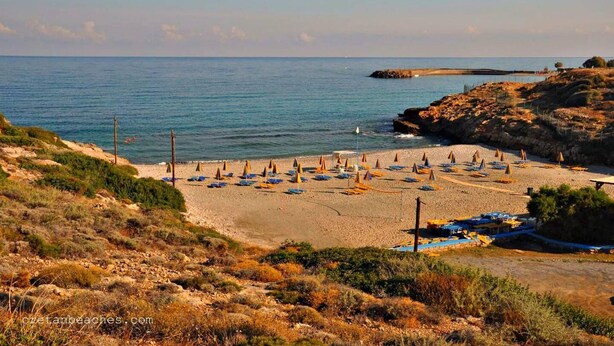A beach with rows of umbrellas and lounge chairs, rocky terrain, and a clear view of the sea. - Spotless Apt in the Heart of Sisi (Agios Nikolaos)