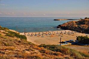 A beach with rows of umbrellas and lounge chairs, rocky terrain, and a clear view of the sea. - Spotless Apt in the Heart of Sisi (Agios Nikolaos)