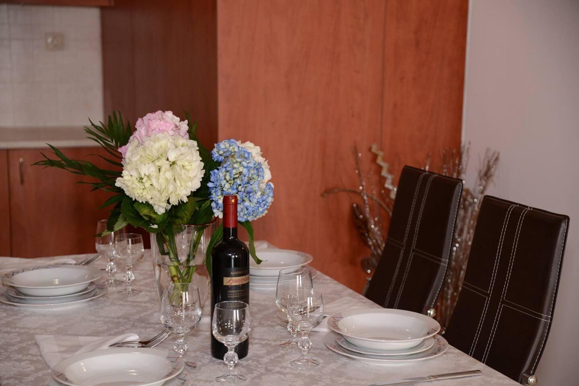 A dining table set with white plates, wine glasses, and a bottle of wine. There are two floral arrangements, one with white flowers and another with blue flowers.