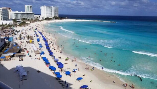 Ein Strand mit einer Reihe blauer Sonnenschirme und Liegestühlen, ein sandiger Uferstreifen und Gebäude im Hintergrund.