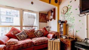 A cozy living room with a red sofa, patterned cushions, a wooden coffee table, a bookshelf, and a wall clock. - Amazing Riverside Boheme Home in SW London! (London)